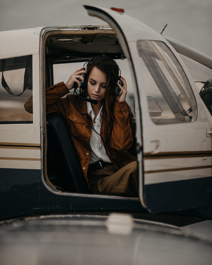 Charming female with long hair in trendy wear and headset sitting in cockpit of modern airplane with opened door in airfield